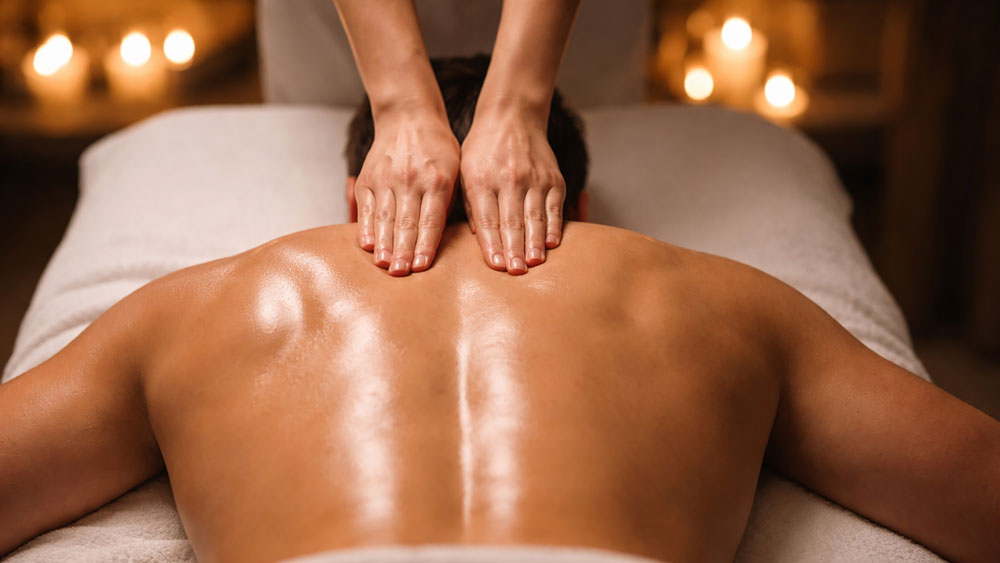 Swedish massage in Belgrade hotel room, female therapist performing diagonal gliding strokes on male side body and lower back with warm ambient lighting